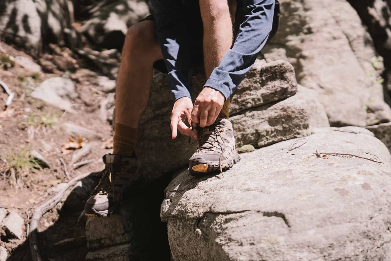 A hiker sitting on a boulder, tying the laces of a hiking boot